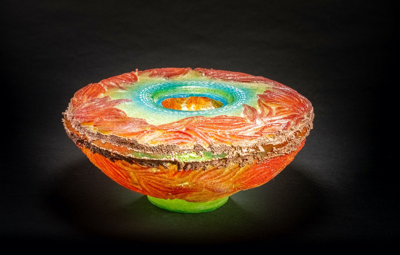 Colorful glass bowl, vibrant orange and green exterior, with blue and orange center, set against black backdrop.