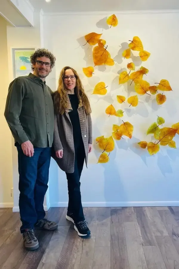 A couple stands by a wall art of yellow leaves. Man wears green shirt, jeans. Woman wears brown coat, black pants.