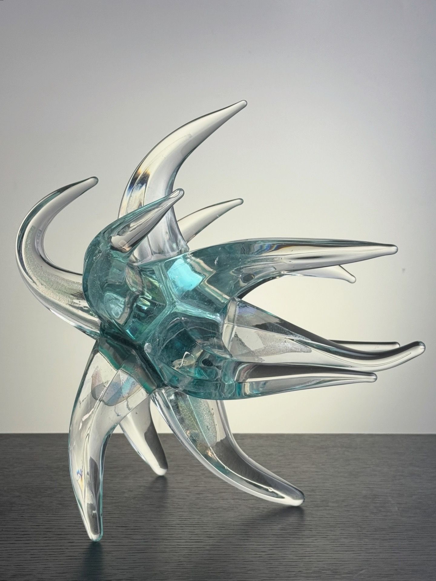 Glass sculpture, turquoise and clear, radiating spiky arms, on a dark surface, neutral backdrop.