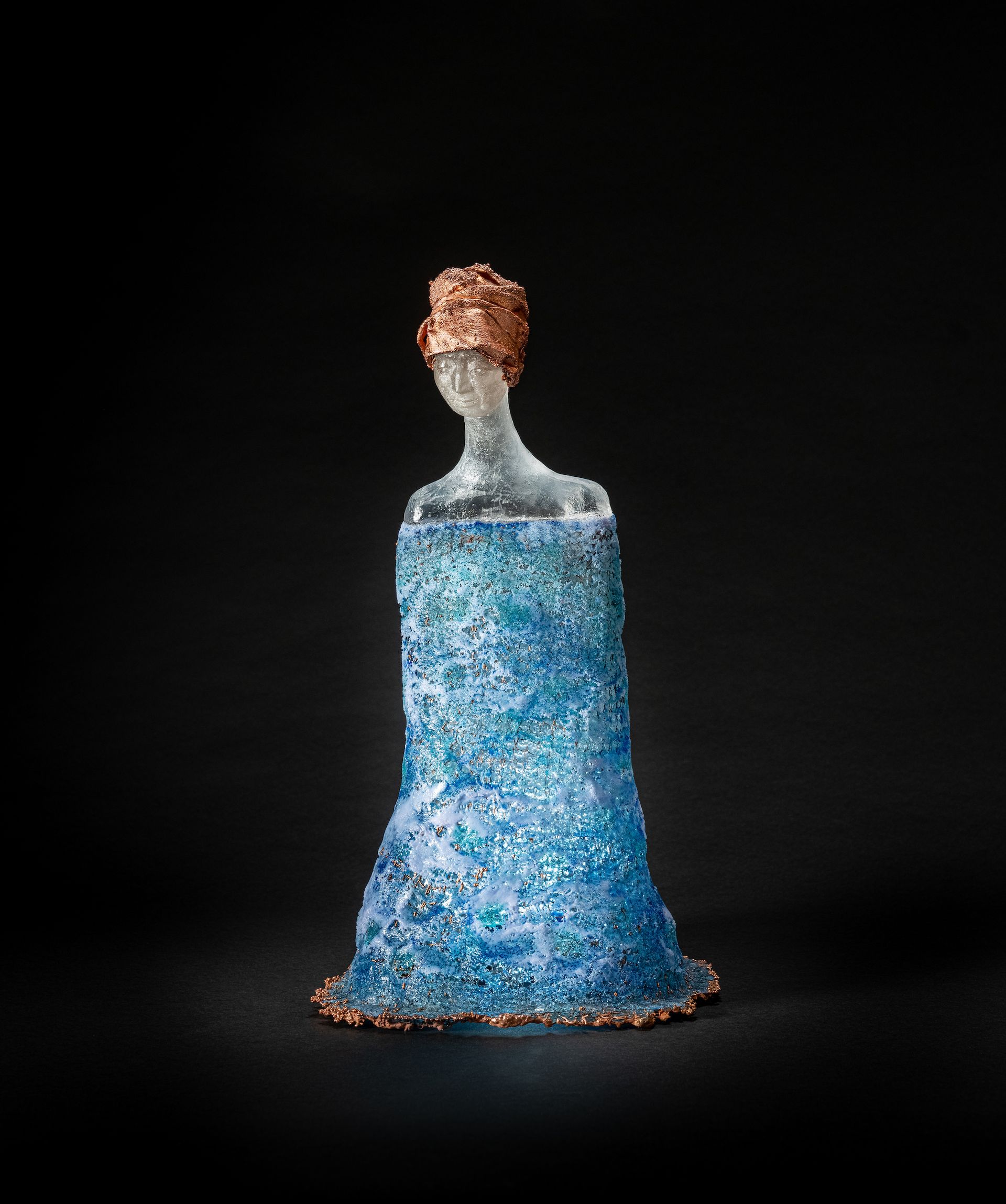 Glass sculpture of a figure in a blue dress and copper-colored headpiece, against a black background.