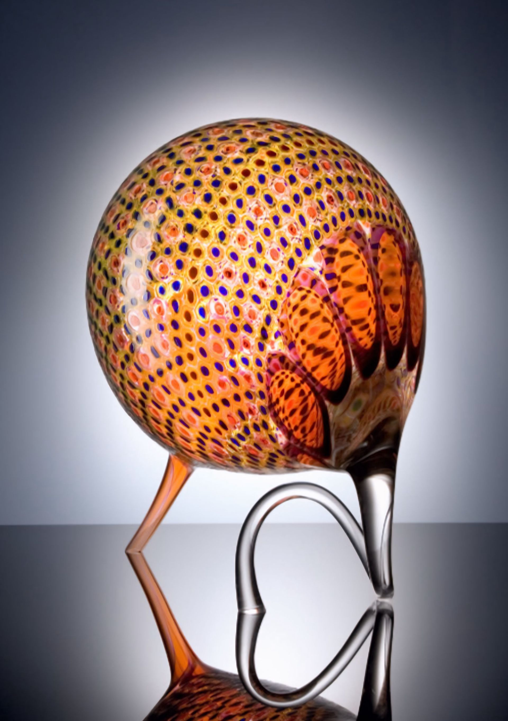 Glass sculpture of an armadillo with a speckled orange body, perched on curved legs, reflected on a dark surface.