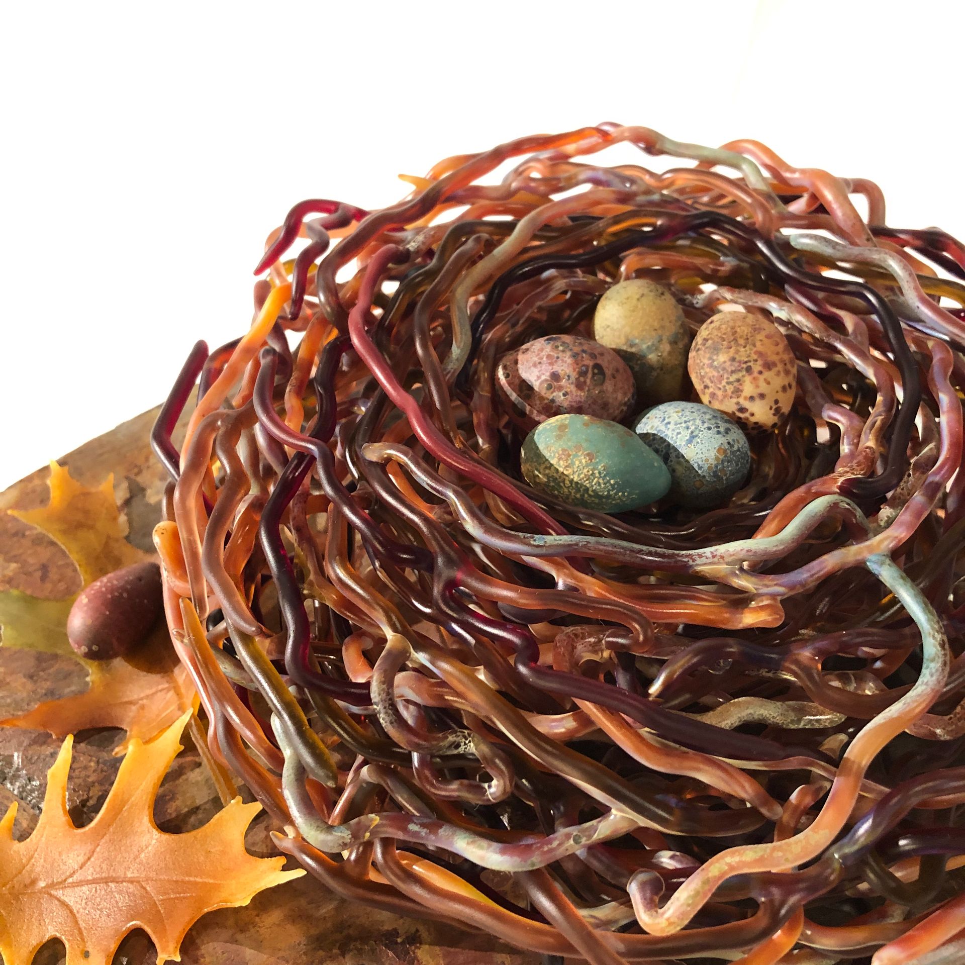 Bird's nest made of brown and orange glass strands with five speckled eggs inside, and an oak leaf.