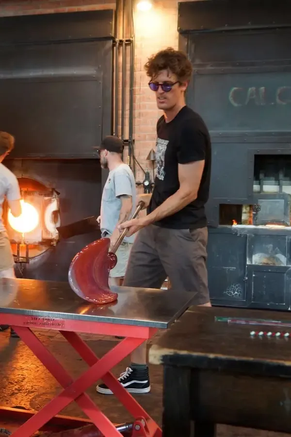 Man working with molten glass near furnace, holding tool, in glassblowing studio.
