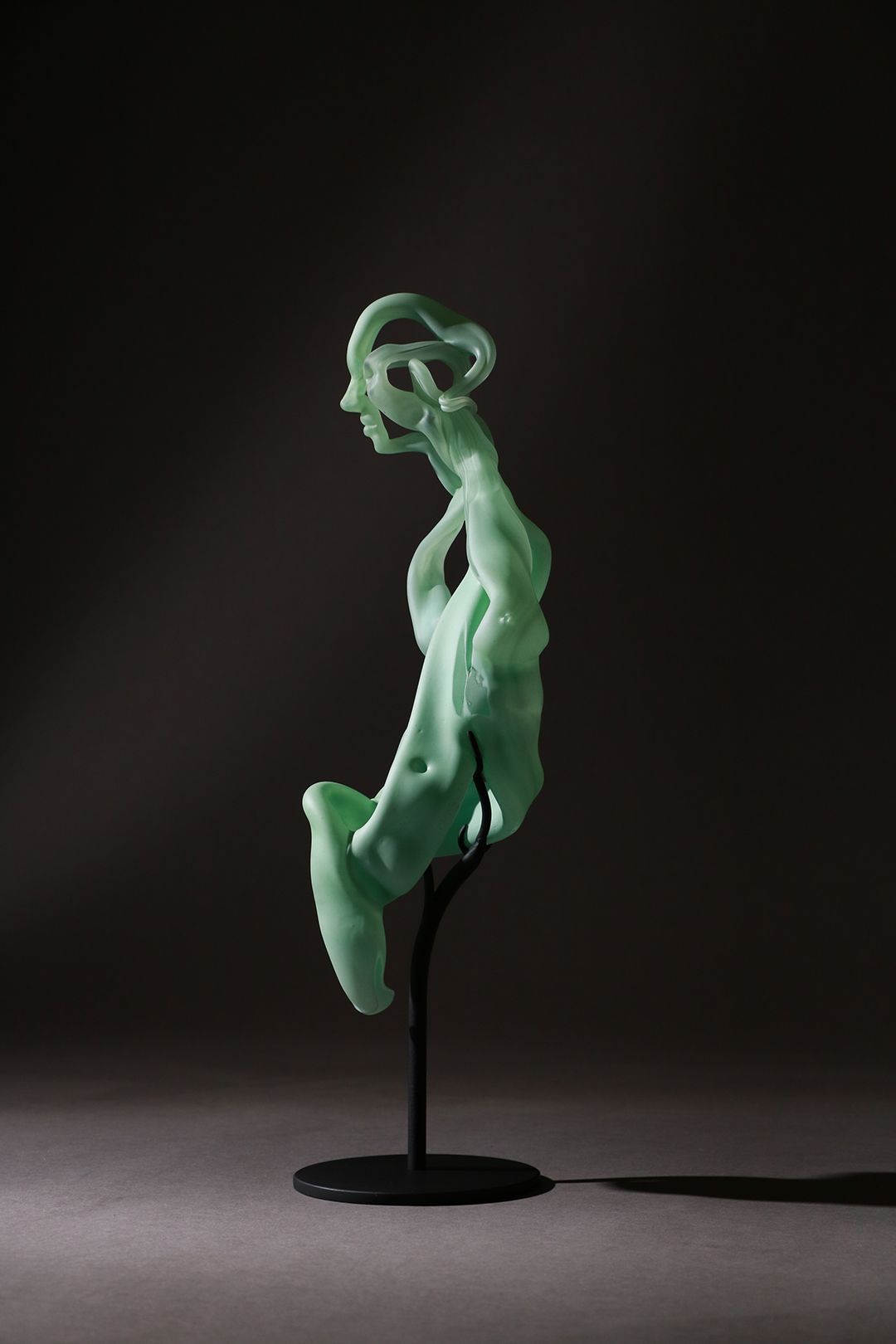 A pale green abstract sculpture of a seated figure on a black stand against a dark background.
