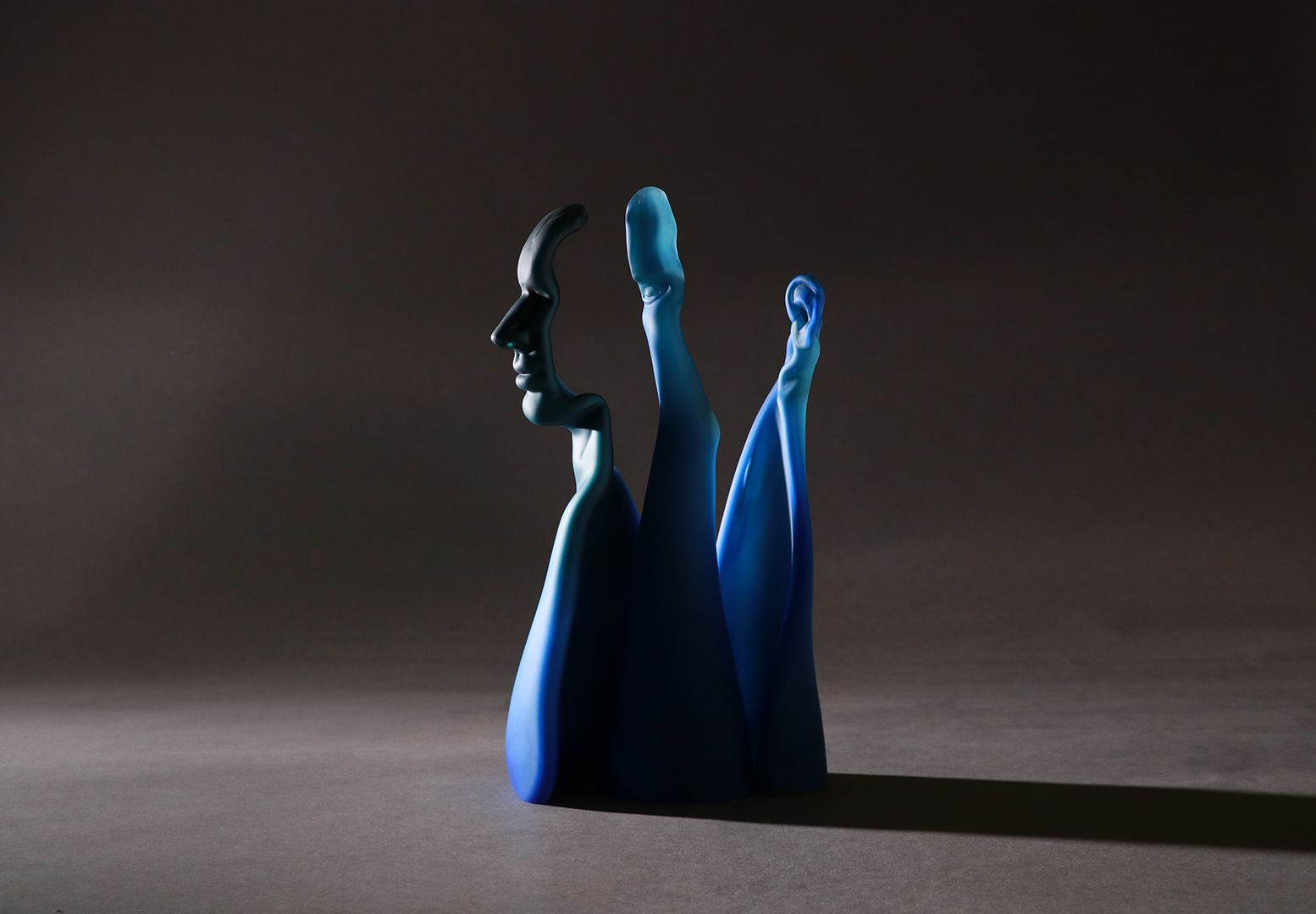 Blue abstract sculpture, face profile on top, legs extending upward, casting a shadow on a grey surface.