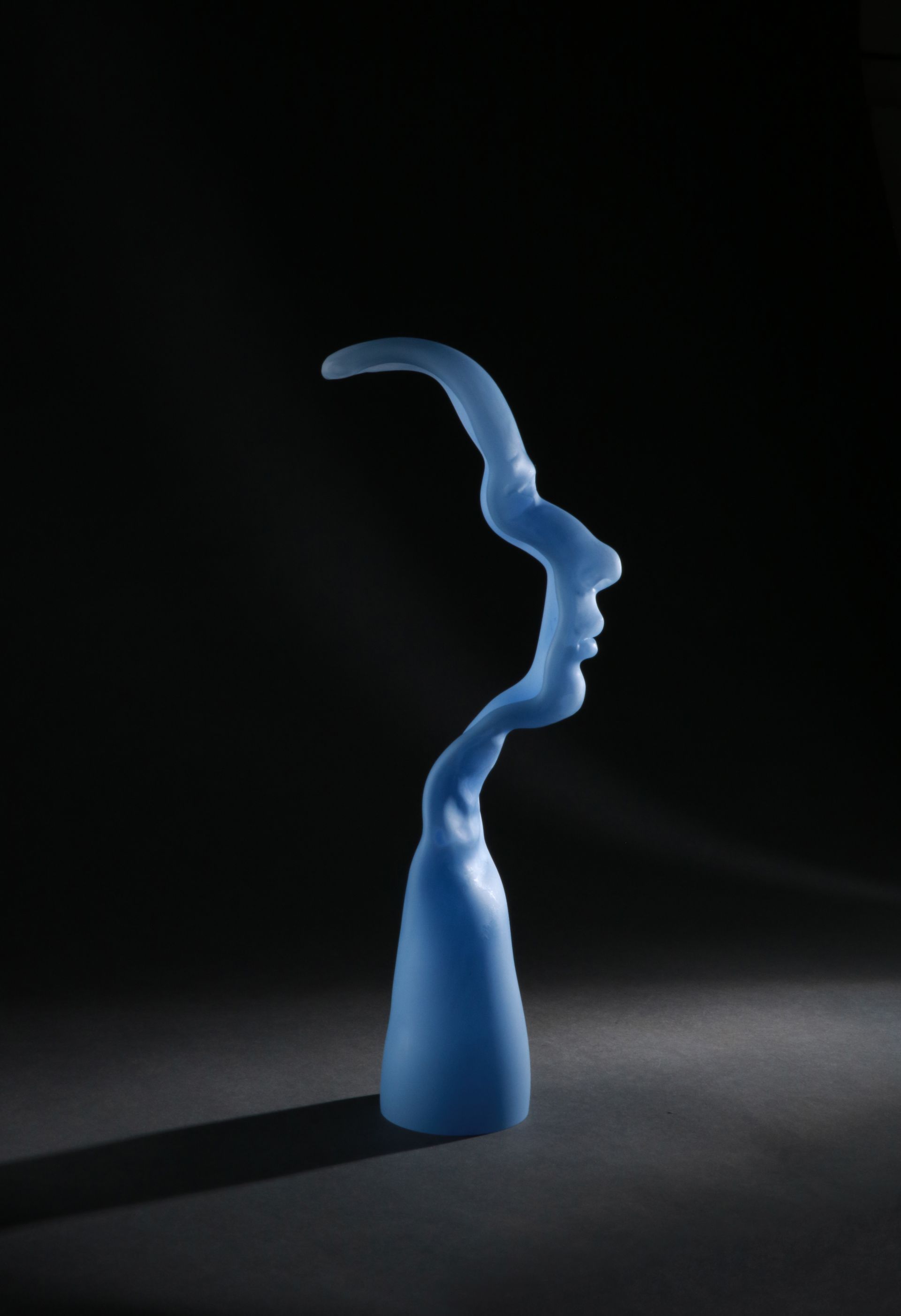 Blue, abstract sculpture of a profile. Vertical, fluid form against a dark background, highlighted.