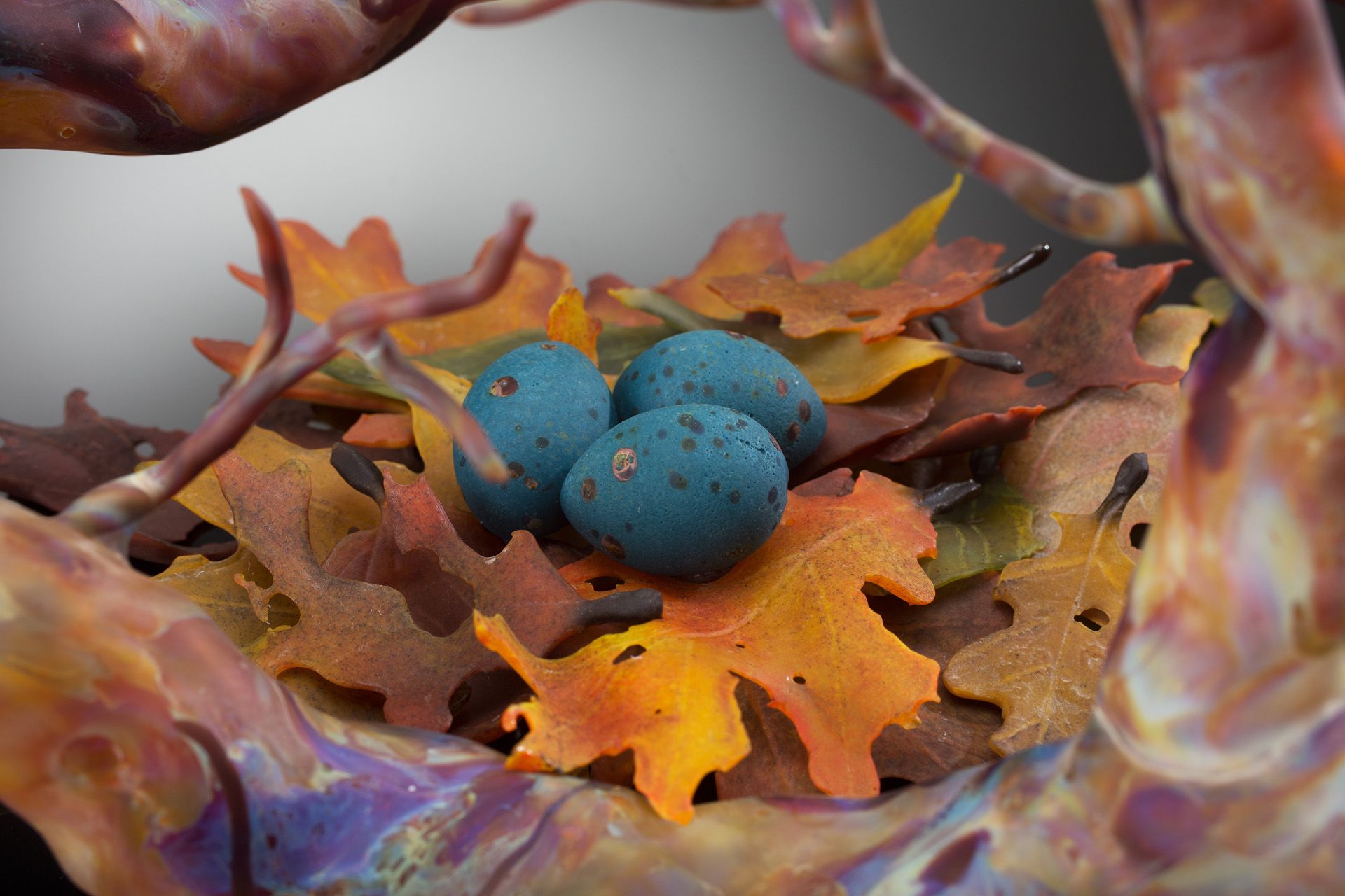 Three speckled blue eggs in a nest of colorful autumn leaves, surrounded by branches.