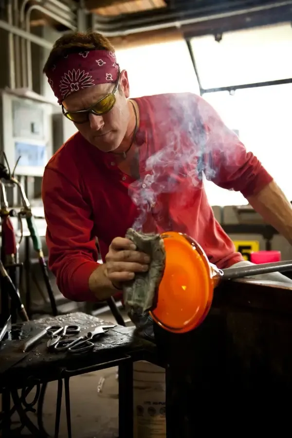 Glassblower shaping molten orange glass with a wet cloth in a workshop.