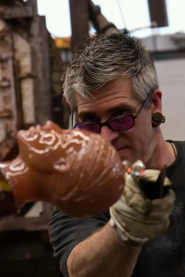 Man shaping molten glass head in a workshop, wearing safety glasses and gloves, with a furnace visible.