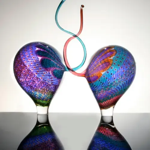 Two iridescent glass bulbs connected by swirling red and blue tubes; reflected on a surface.