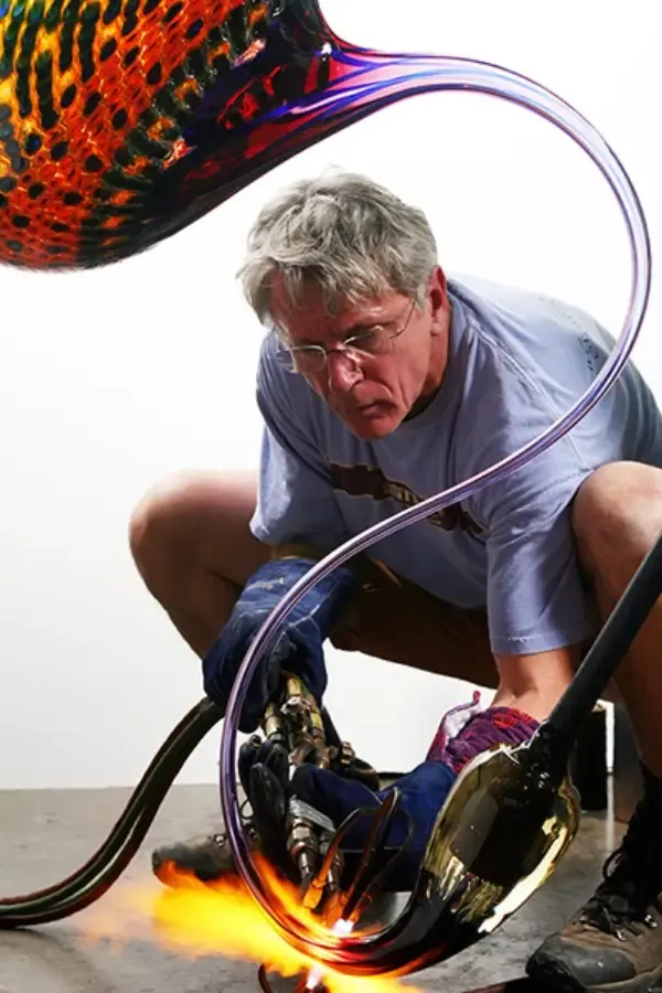 Man using a torch to shape colorful glass sculpture, crouching in a studio with bright orange flames.