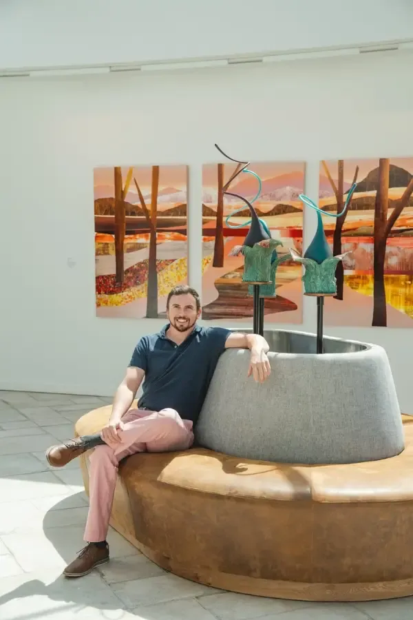 Man sitting by sculpture, wearing pink pants and blue shirt, art in the background, light-filled room.