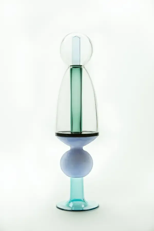 Colorful glass sculpture with teal, blue, and clear sections. Tall, abstract form against a white backdrop.