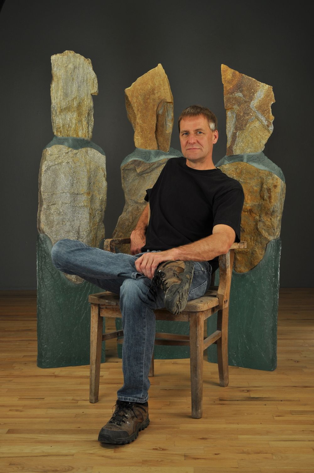 Man sitting in a chair, posing in front of abstract sculptures. He is wearing a black shirt and jeans.