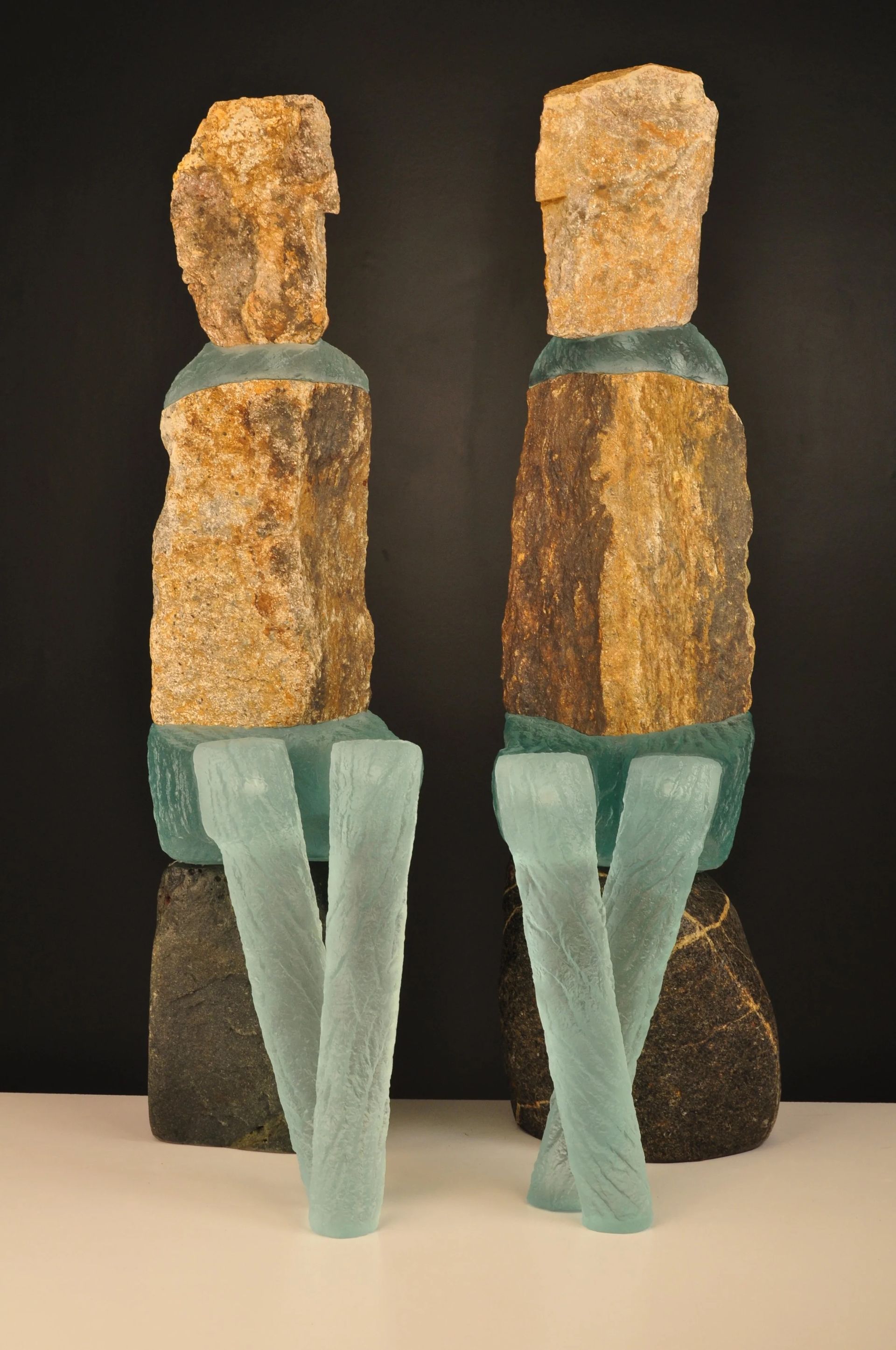 Two abstract sculptures. Rough brown stone heads and torsos atop clear blue glass legs. Dark background.