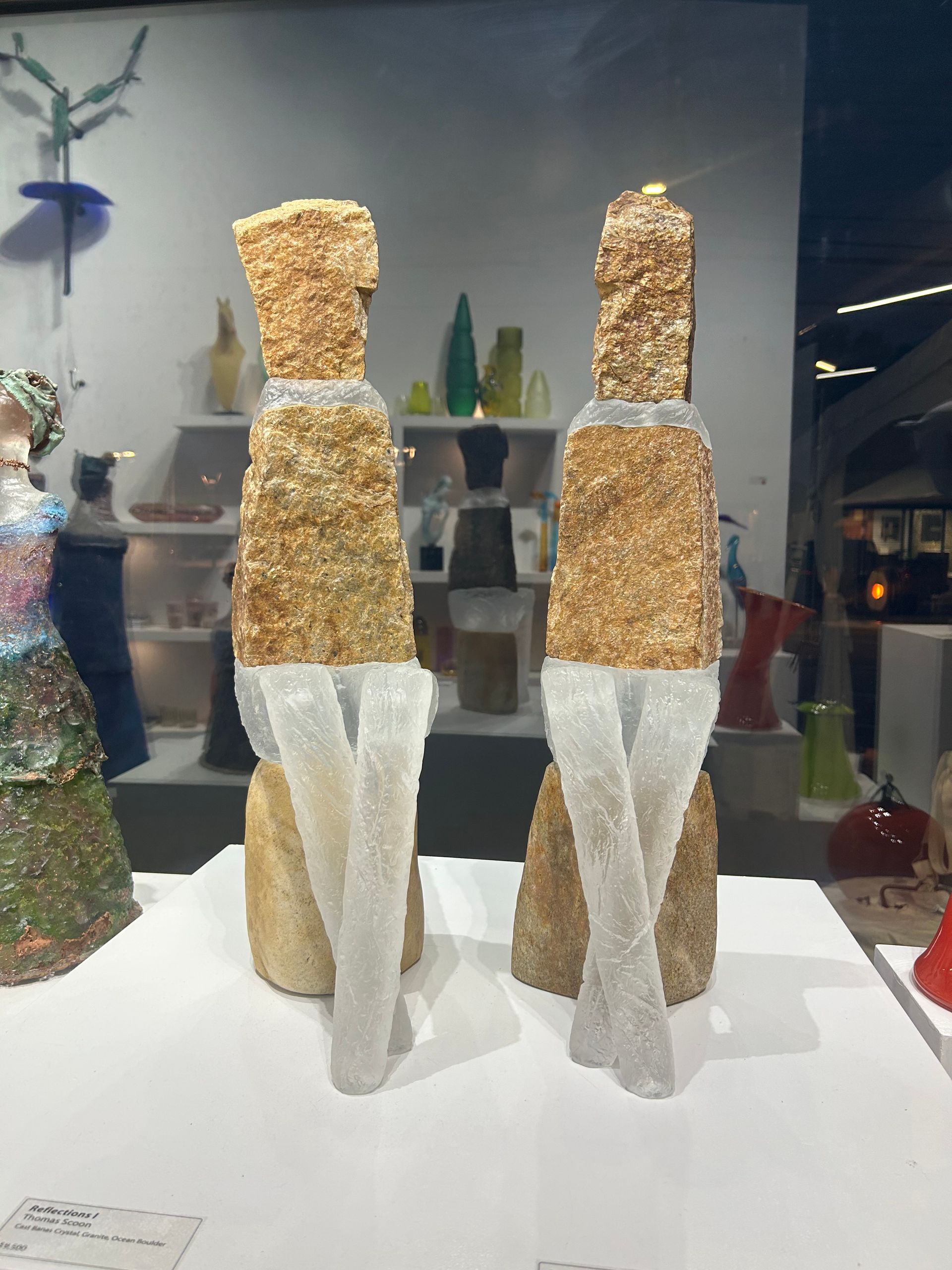 Two sculptures of figures in a display case. Rough, tan torso forms with clear, glass legs crossed at the ankles.