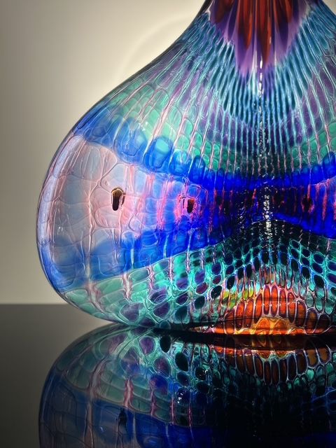 Colorful, blown glass art with blue, teal, purple, and orange hues; reflected on a black surface.