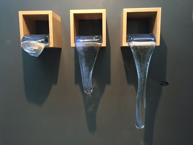 Three glass sculptures resembling water pouring from wooden boxes, mounted on a dark wall.
