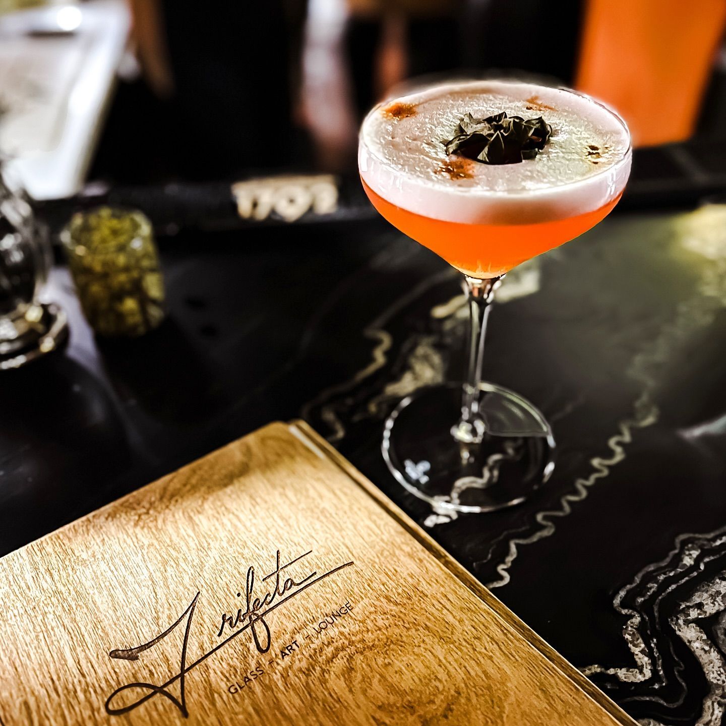 Cocktail on a bar with menu. Orange drink with foam and garnish, on a black table.