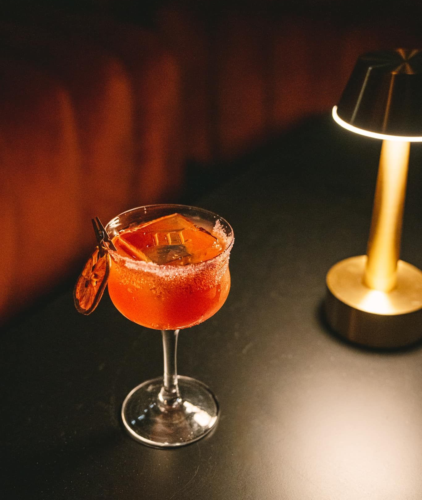 Cocktail in a stemmed glass with orange slice garnish, beside a small lit lamp on a dark surface.