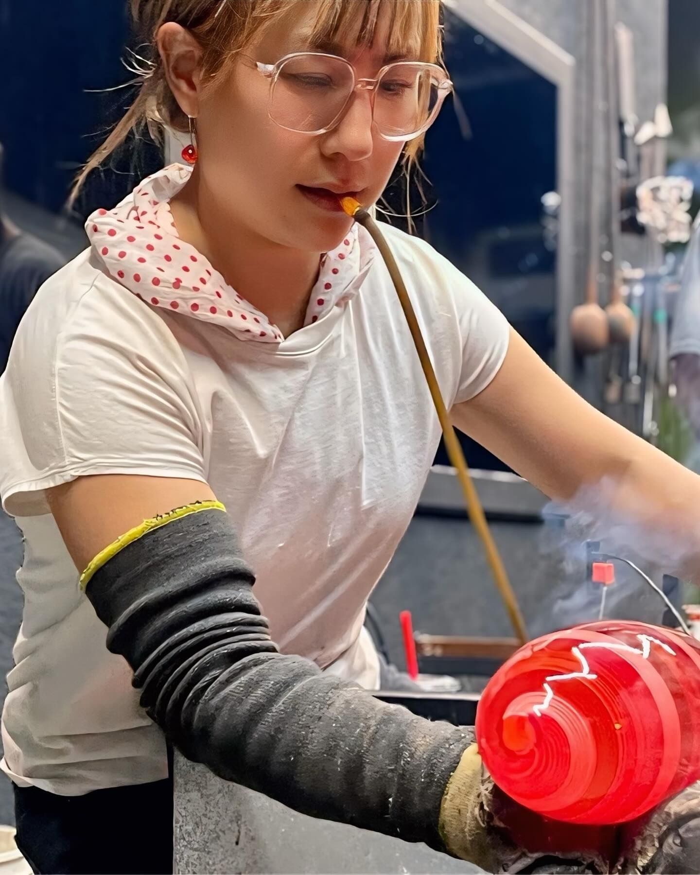 Woman wearing safety gear, blowing glass with a torch. Red and white glass orb in progress.