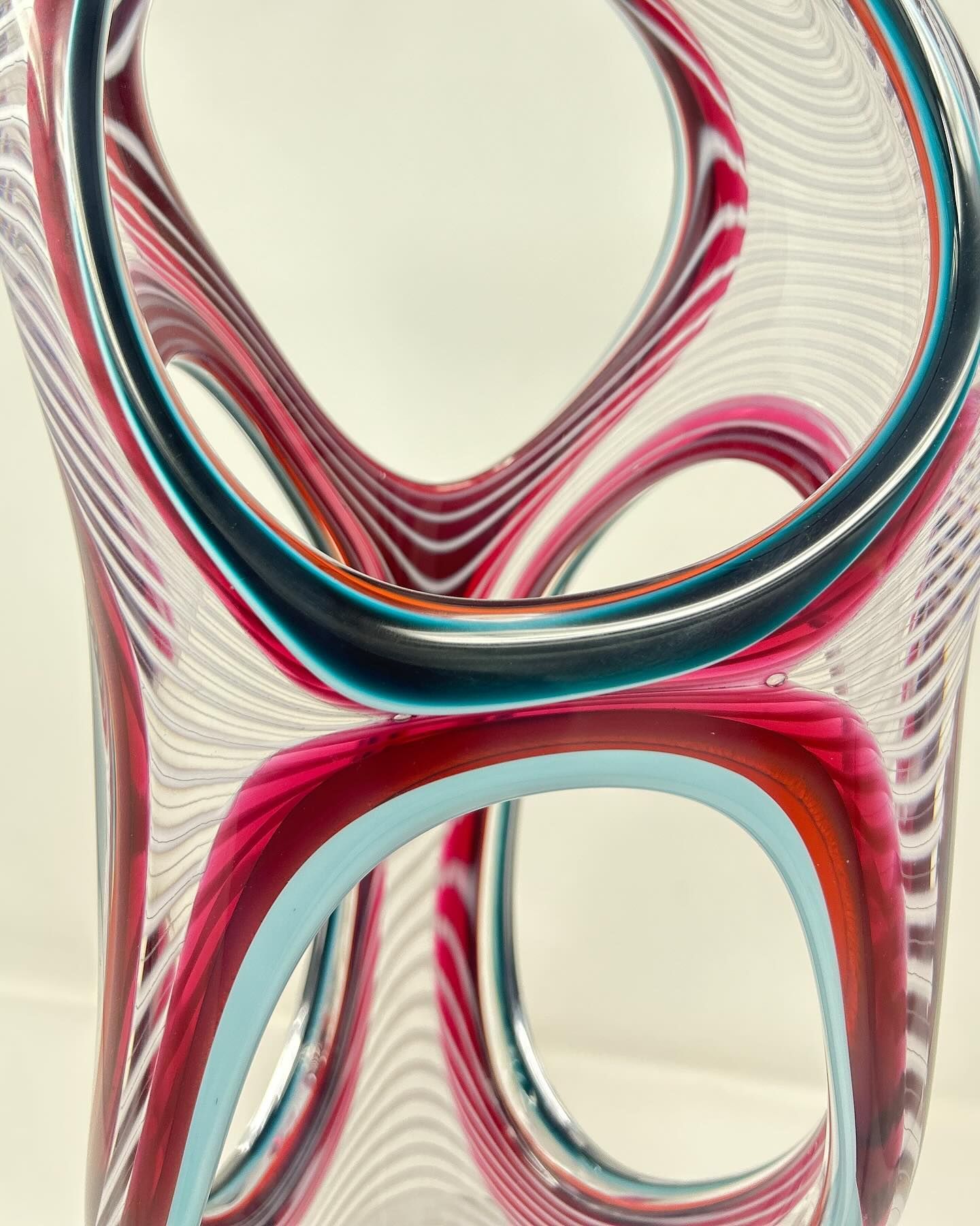 Abstract glass sculpture with intertwined red, teal, and clear stripes.