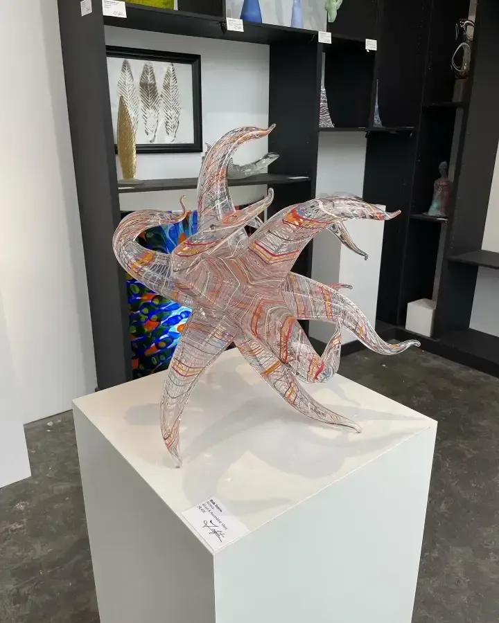 Glass sculpture on white pedestal in gallery setting; red and clear with abstract shapes.