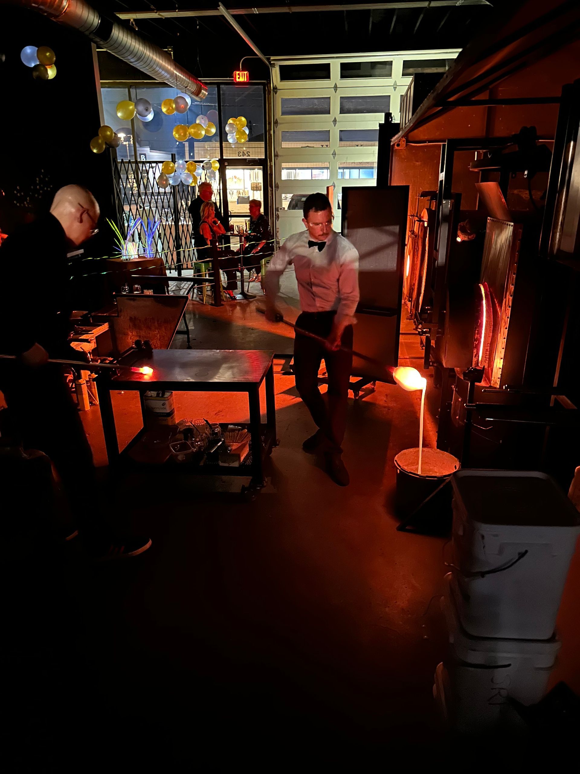 Glassblower at work in a dimly lit studio. He’s shaping molten glass on a table near a furnace.