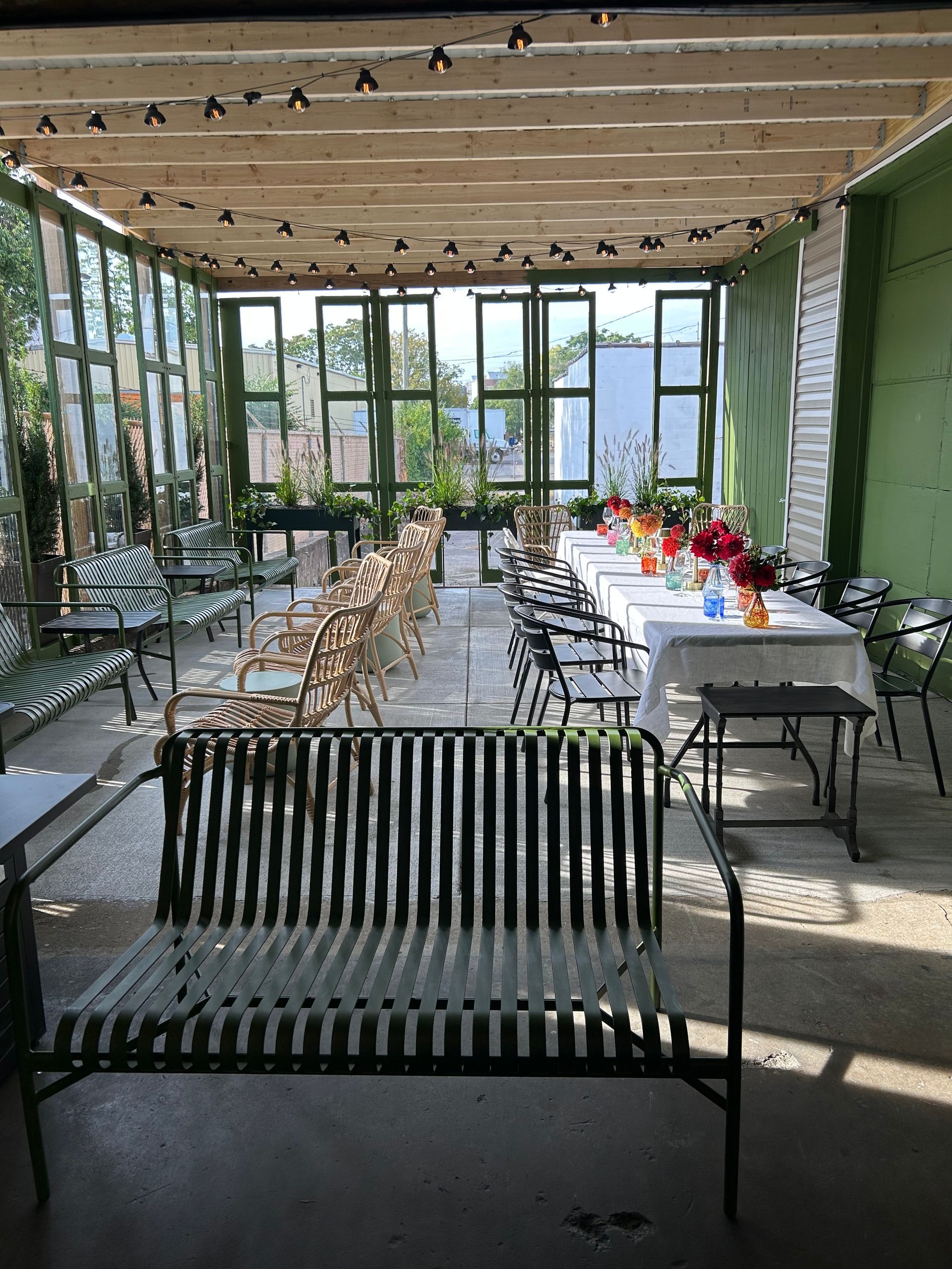 Outdoor dining area with tables set for a meal, rows of chairs, bench, green walls, string lights.