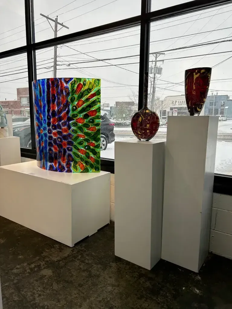 Glass art on white pedestals in a gallery setting with a window view of an overcast day.