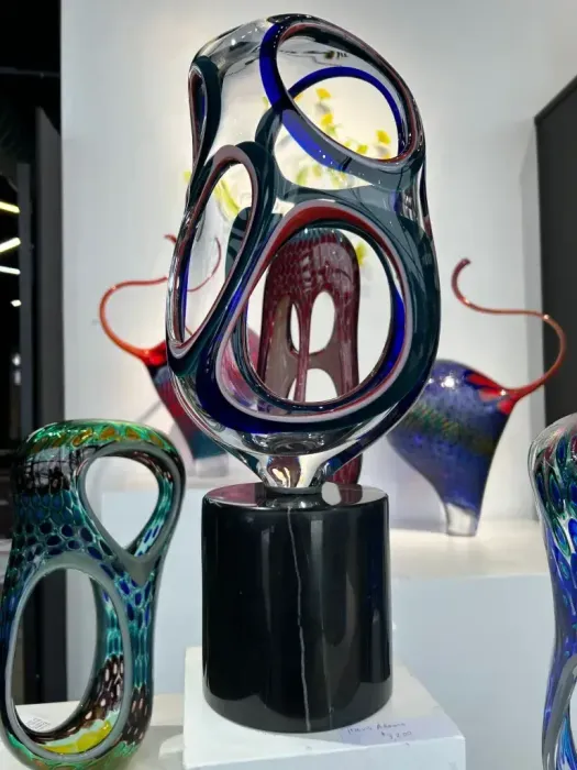 A tall, abstract glass sculpture with blue and red accents, on a black base, displayed in a gallery.