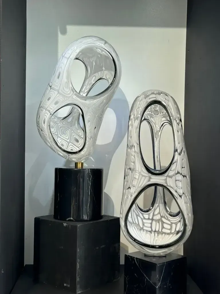 Two abstract glass sculptures with carved designs, atop black pedestals.