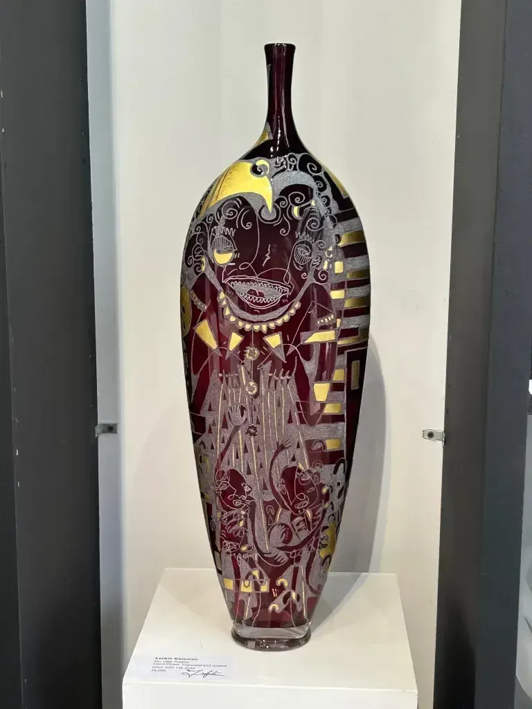 Tall, burgundy glass vase with intricate gold and silver etched designs, displayed on a white pedestal.