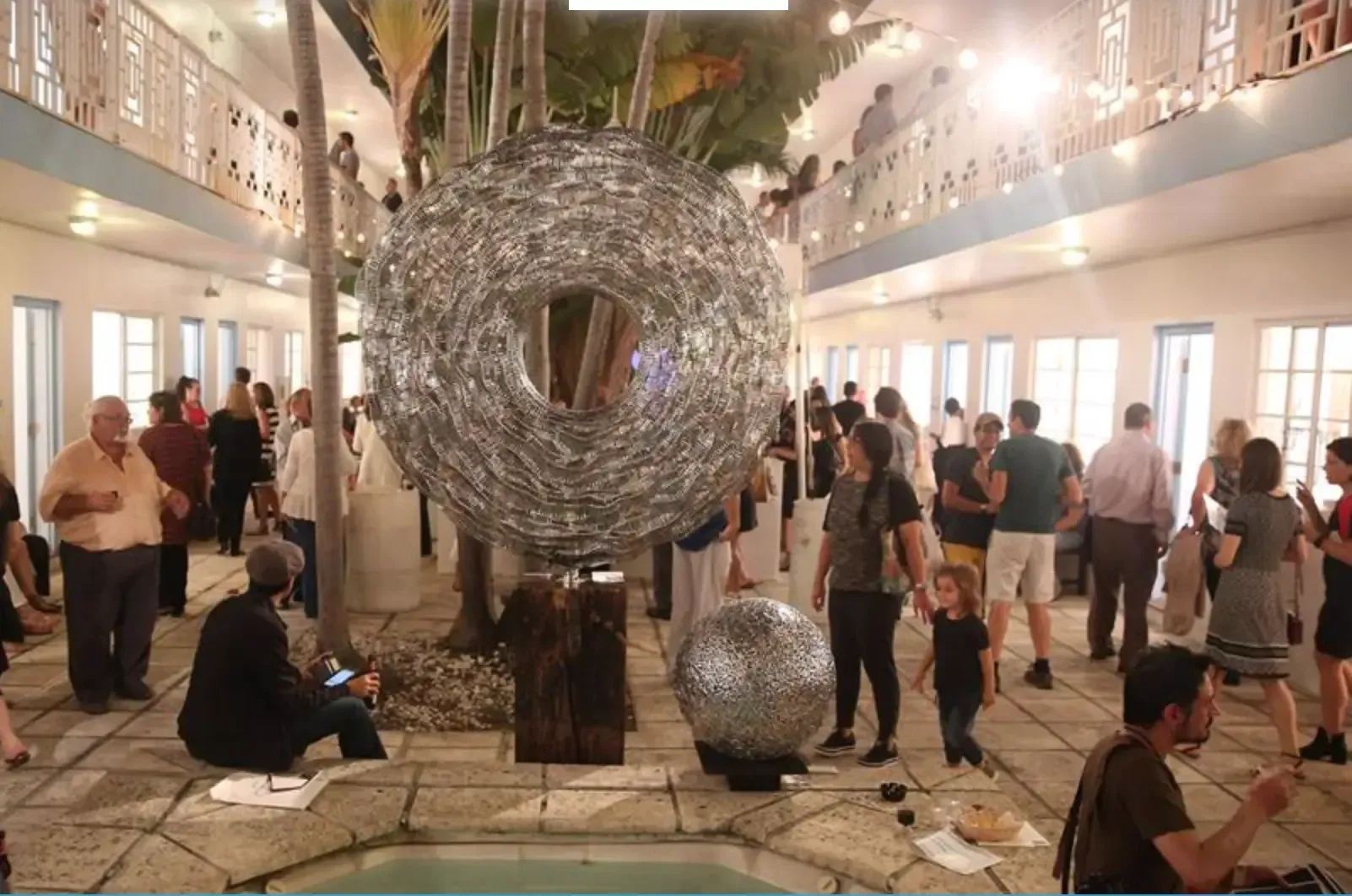 People at art gallery, large metallic sculpture. Courtyard setting with architectural details, trees, and small reflecting pool.