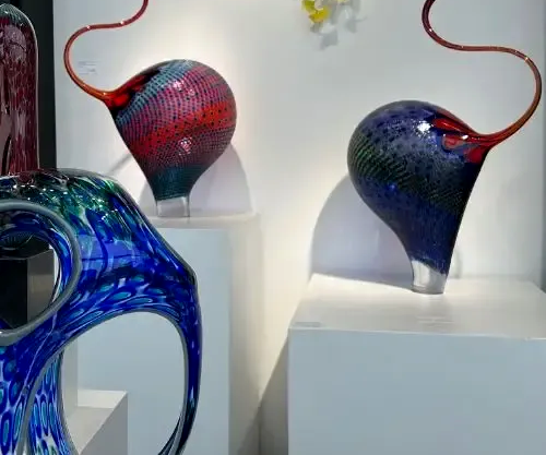 Glass sculptures of various shapes and colors on white pedestals, displayed in a gallery setting.