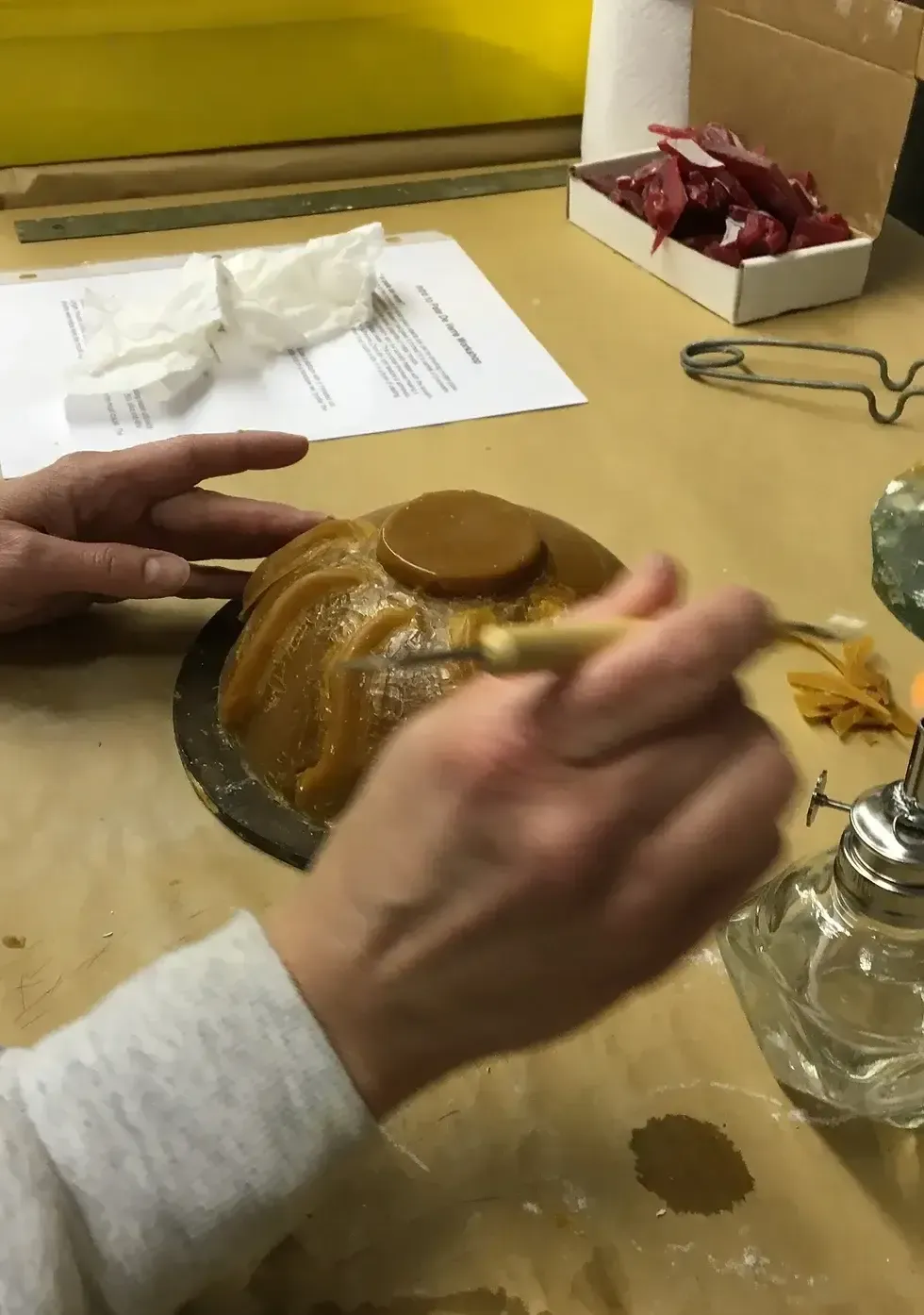 A person melting wax on a mold with a heated tool; work area with materials.