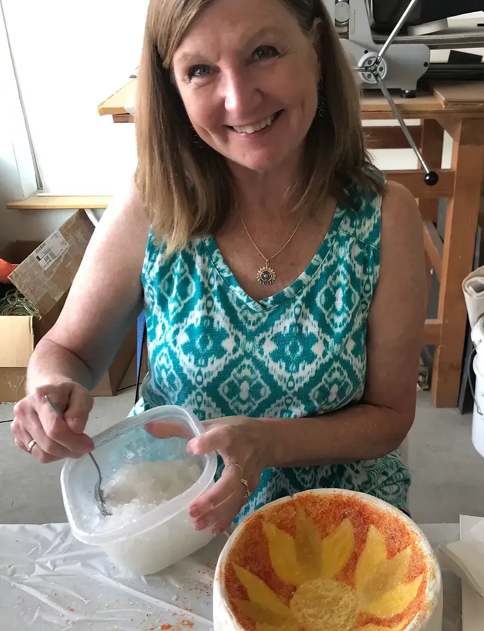Woman stirring a white substance in a container; smiling, sitting next to a sunflower-decorated bowl.