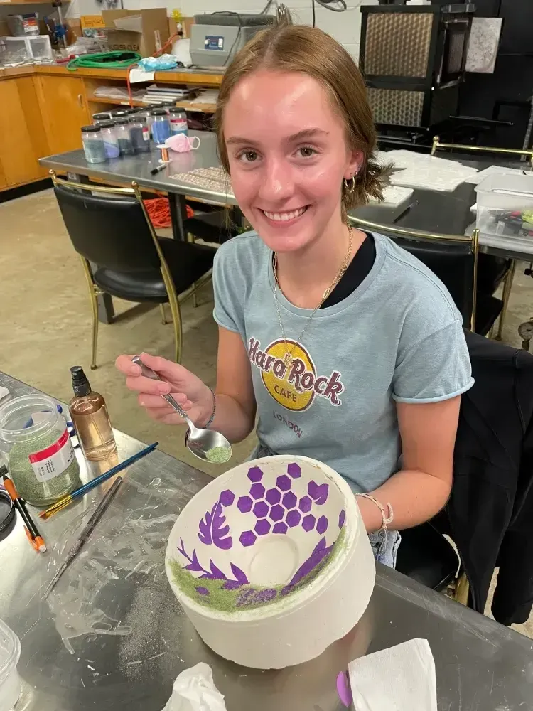 Person smiling, holding a spoon over a decorated bowl. They are in a workshop-like setting with paint supplies.