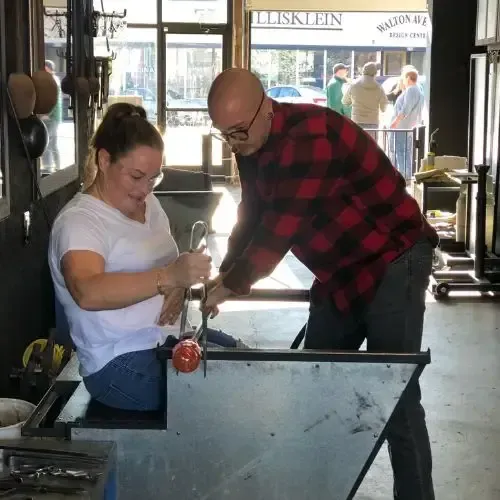 Two people shaping molten glass in a workshop. One holds a tool, the other guides. Interior setting.