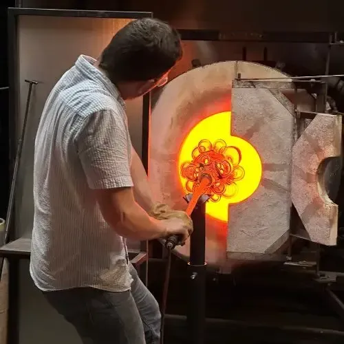 Glassblower, wearing protective gloves, working with molten glass in a furnace, illuminated in orange.