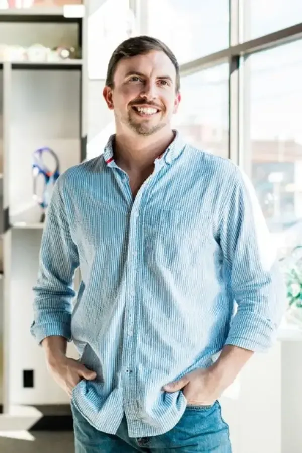 Man in a blue button-down shirt smiles, hands in pockets, near a window.