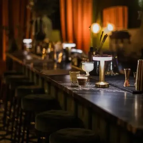 Cocktail bar scene with drinks, stools, and warm lighting.