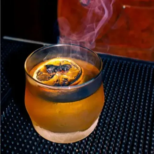 Smoked cocktail in a glass with a charred orange slice. Smoke rises. Set on a black bar surface.