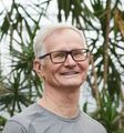Smiling man with glasses in a gray shirt, standing outdoors with leafy plants behind him