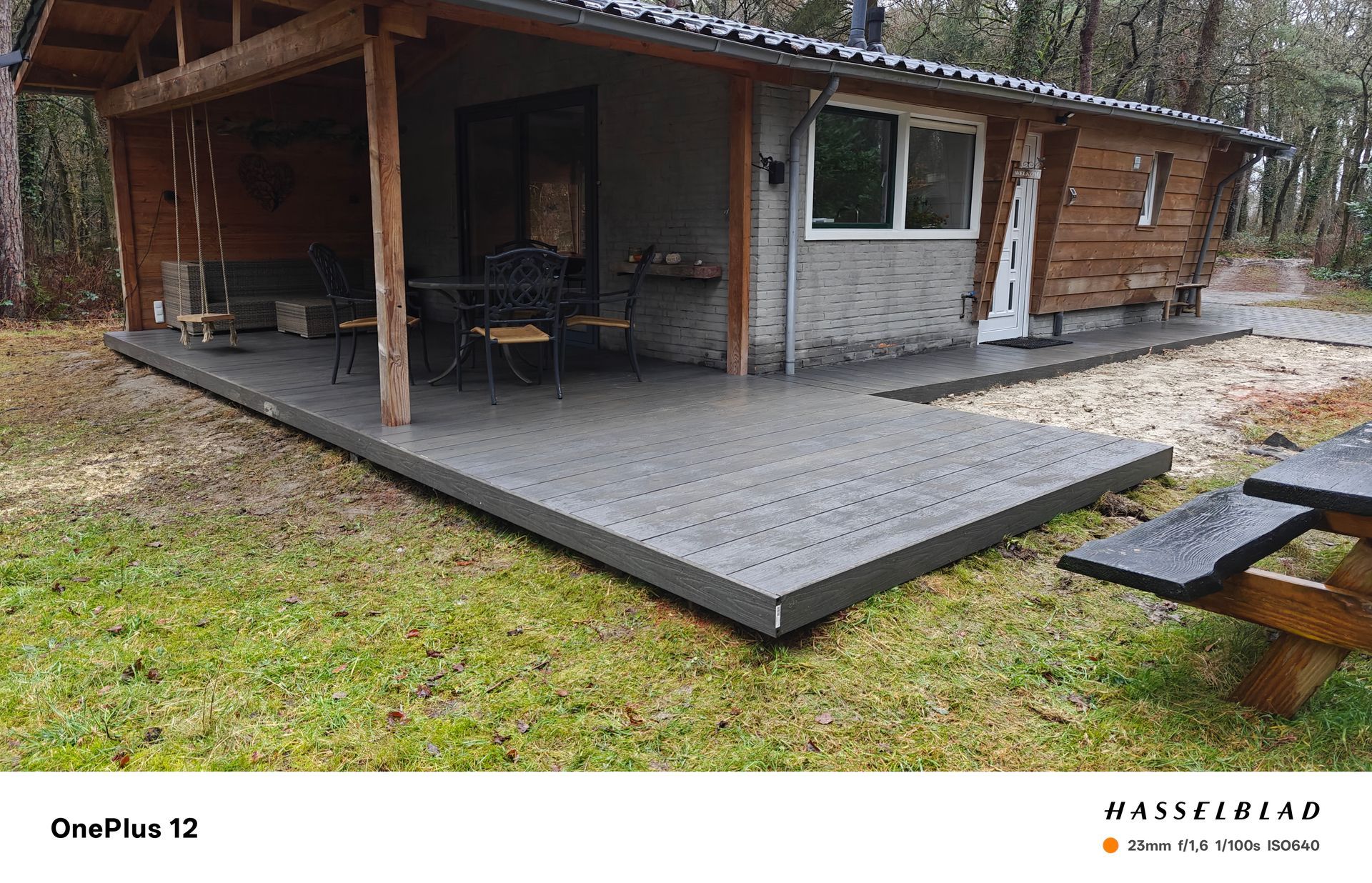 Grijze composiet vlonder voor een kleine blokhut met een veranda; een picknicktafel is zichtbaar in het gras.