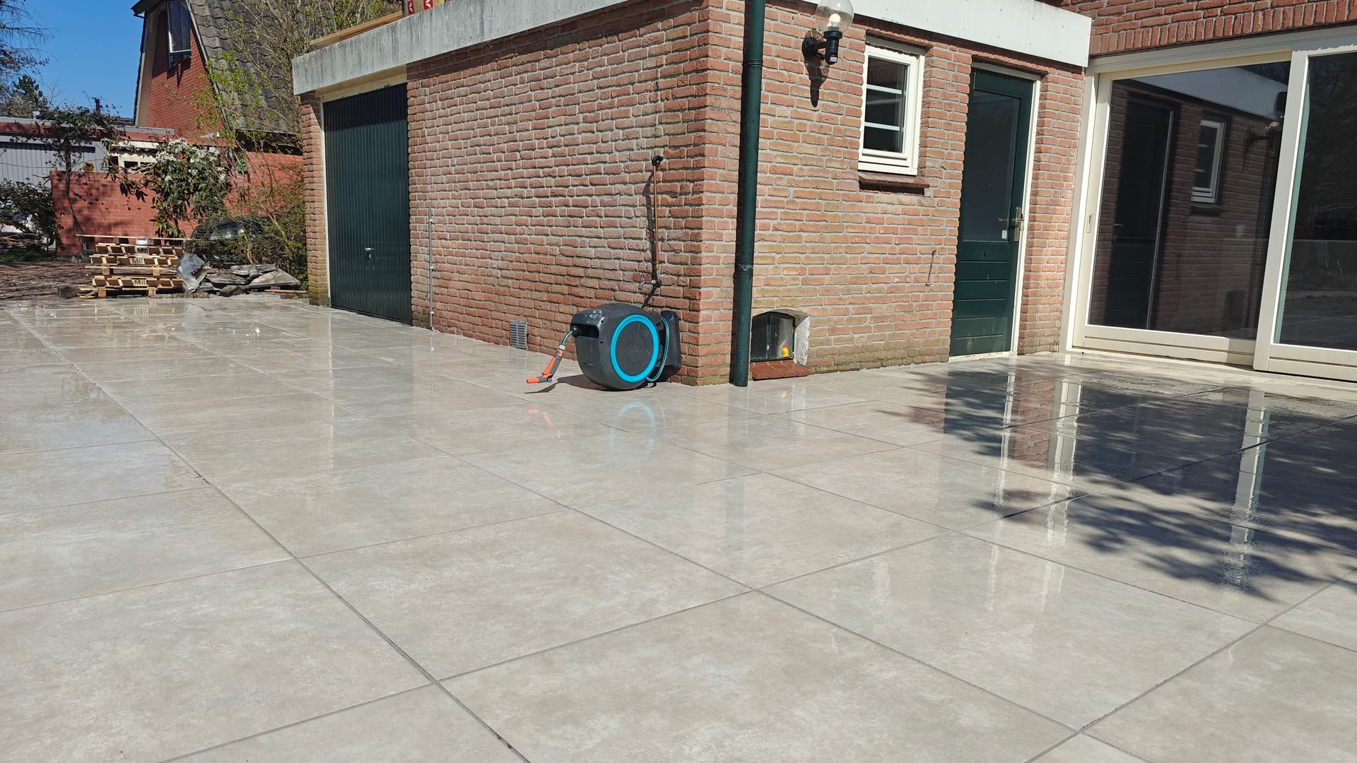 Een glanzend betegeld terras met een bakstenen gebouw en een groene deur. Een tuinslang met haspel staat er vlakbij.