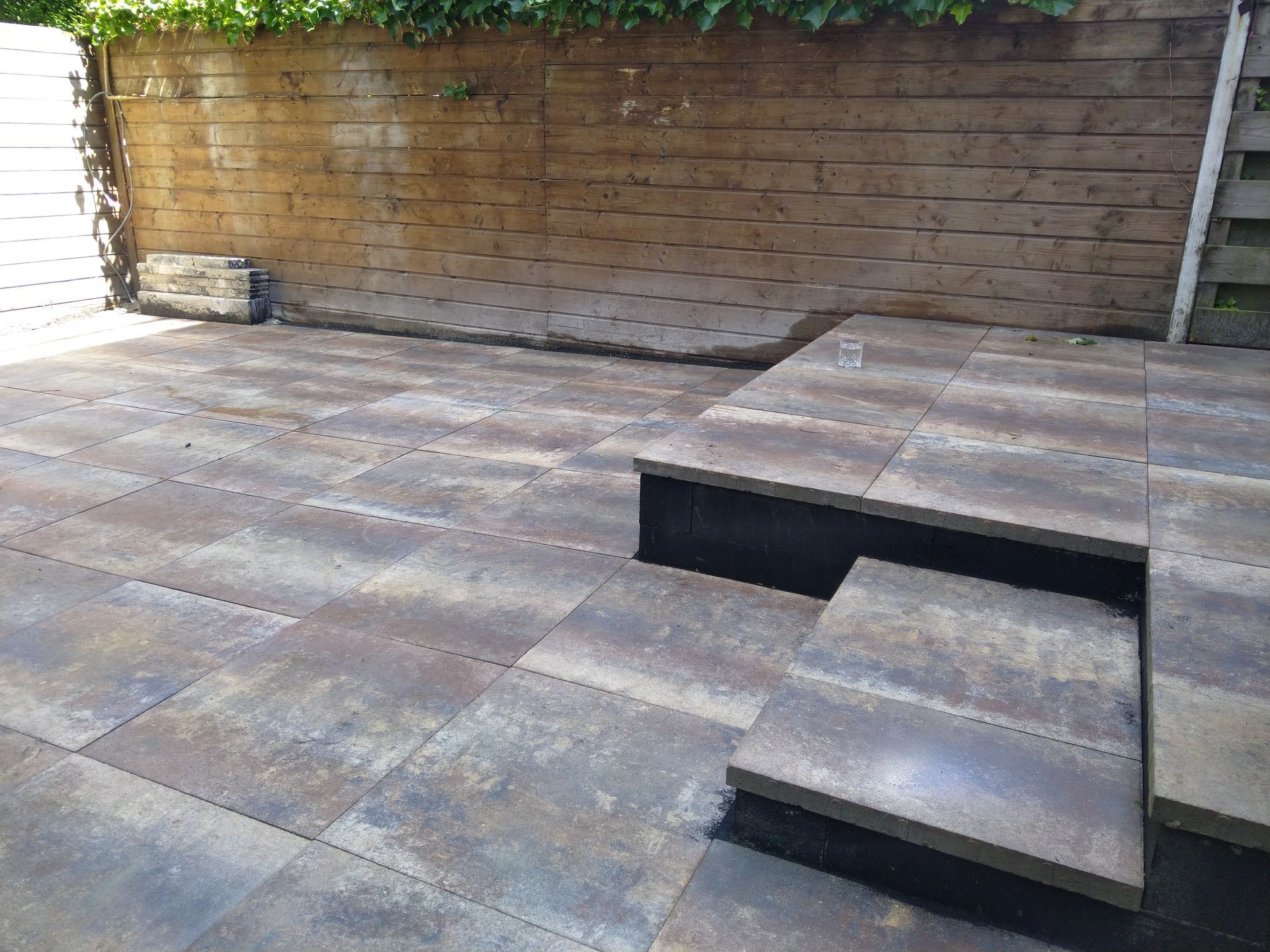 Terras met stenen bestrating en trappen die naar een verhoogd gedeelte leiden. Houten schutting op de achtergrond.