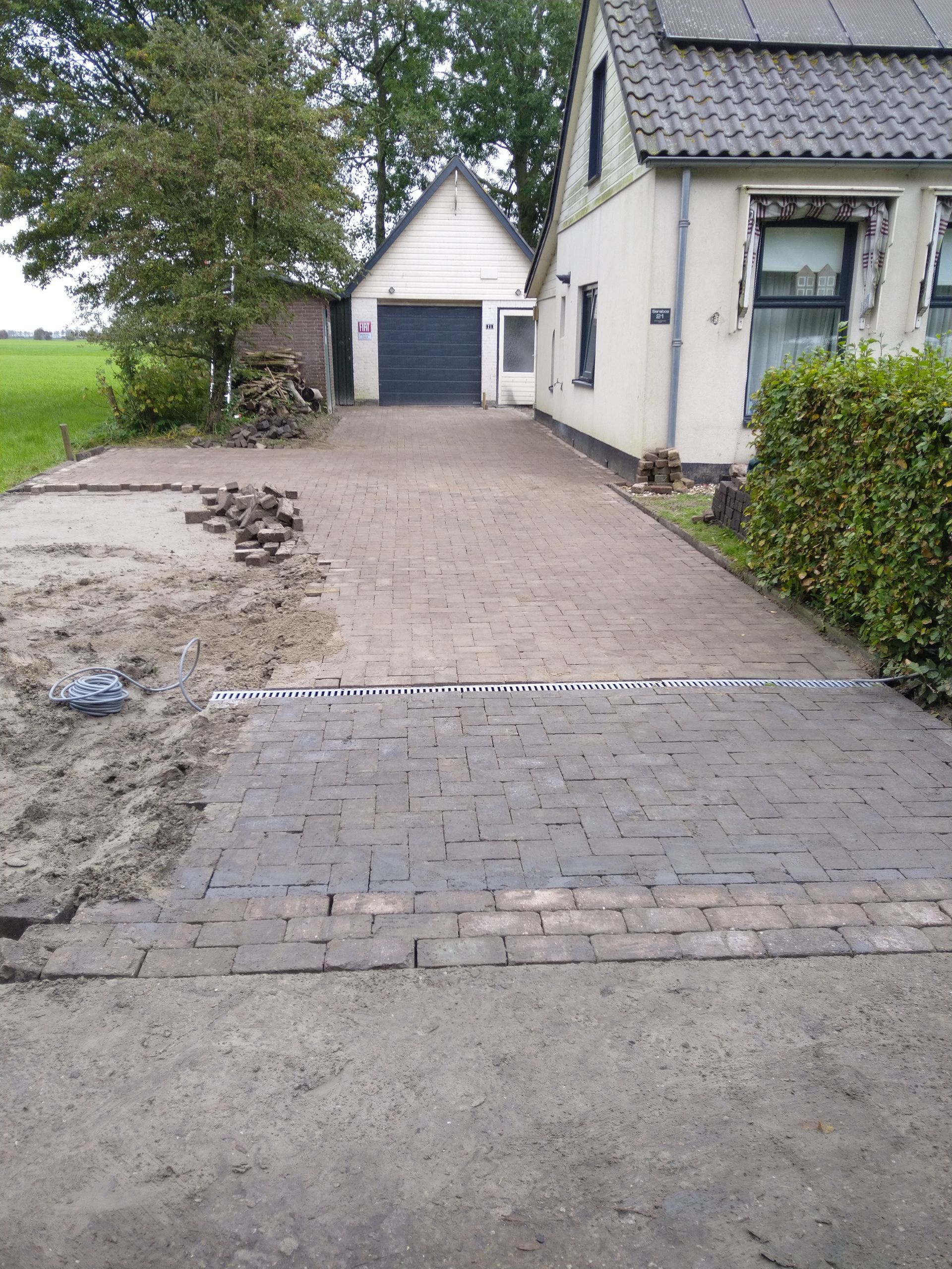 De oprit is gedeeltelijk bestraat met donkere bakstenen en leidt naar een garage en een huis.