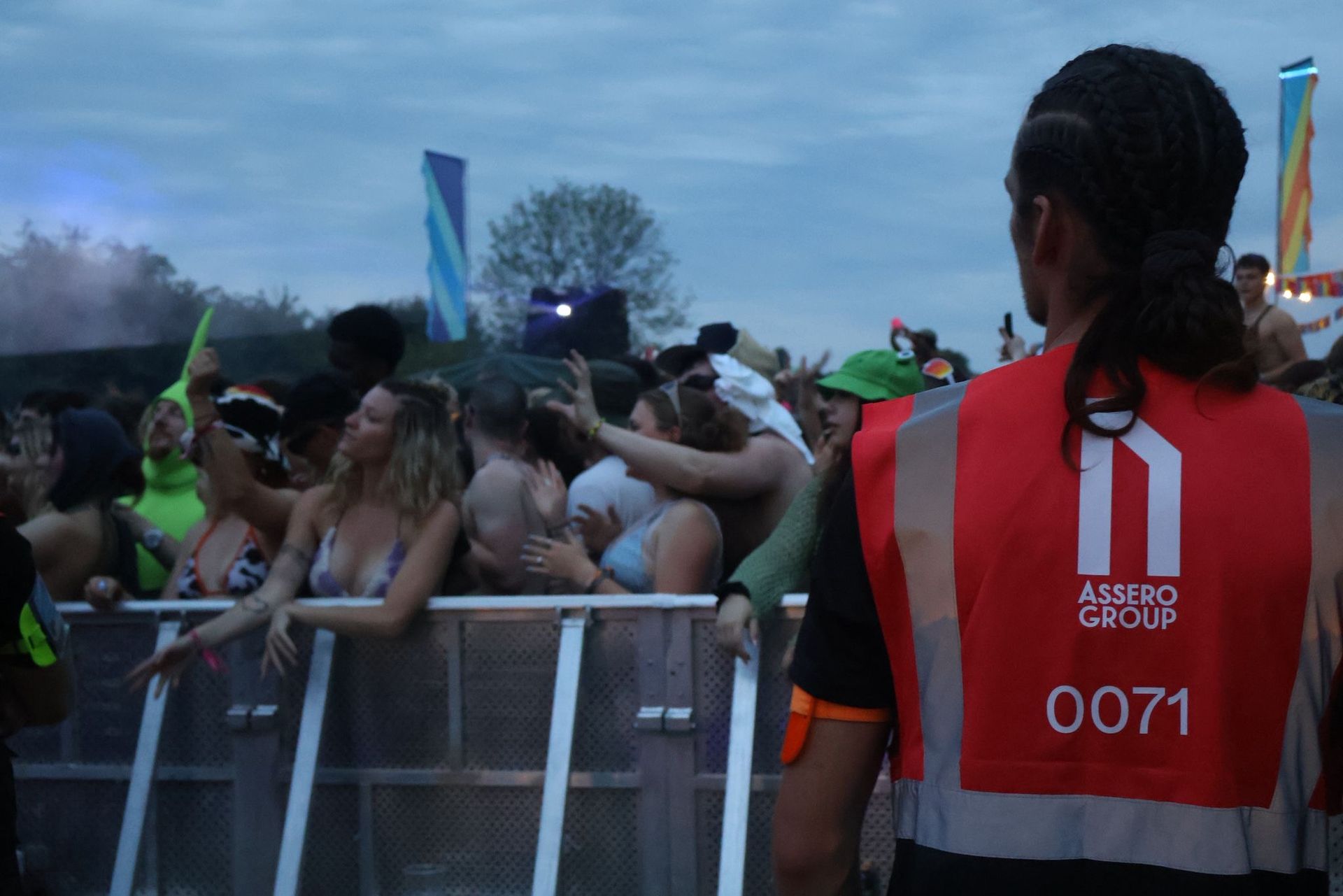 A security guard in a red reflective vest watches a crowd of people standing against a metal barrier at an outdoor event.