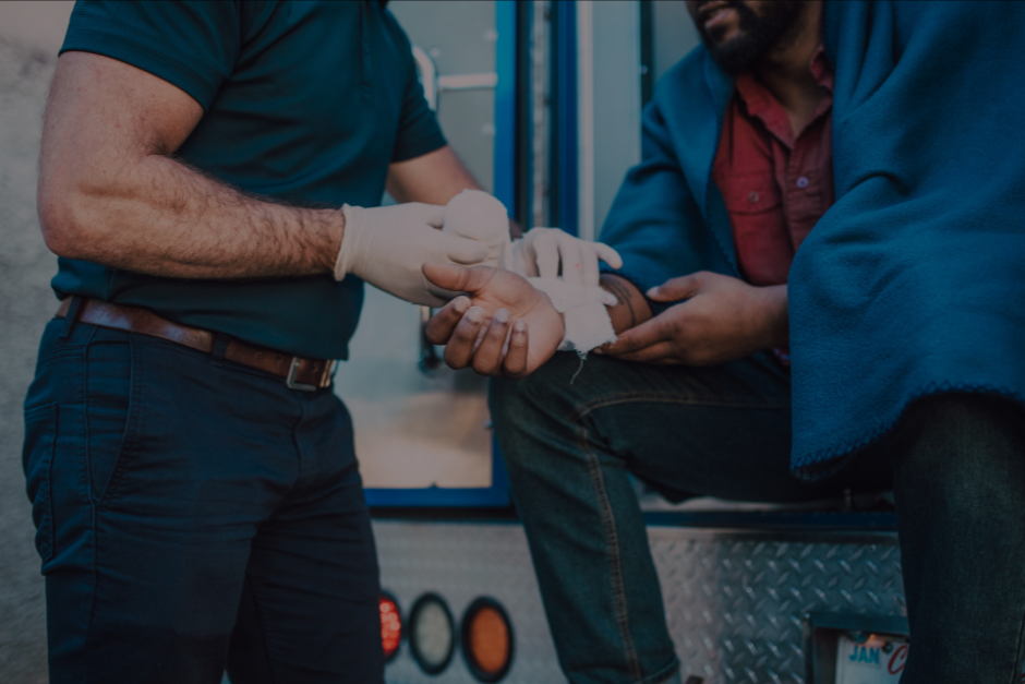Paramedic bandaging a person's hand, outdoors near an ambulance. The person is wrapped in a blanket.